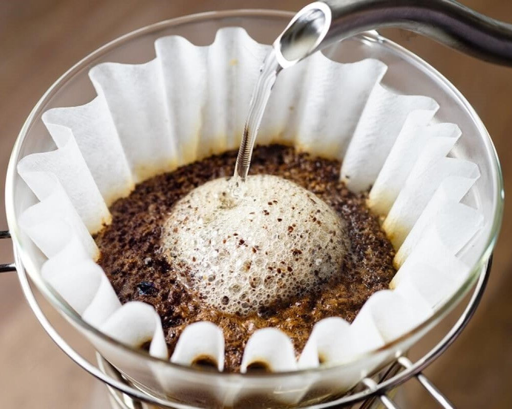 Gooseneck kettle pouring hot water over coffee grounds in a paper filter over a glass pot